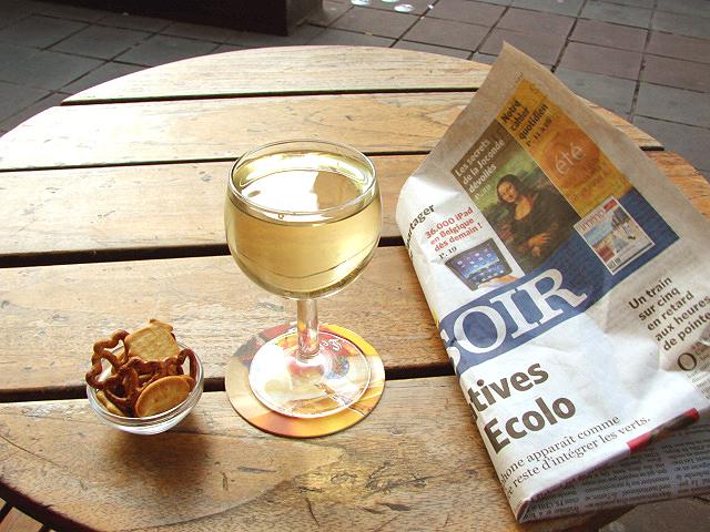 Een tafeltje op een terrasje, met een glas witte wijn, borrel-zoutjes en de krant Le Soir.